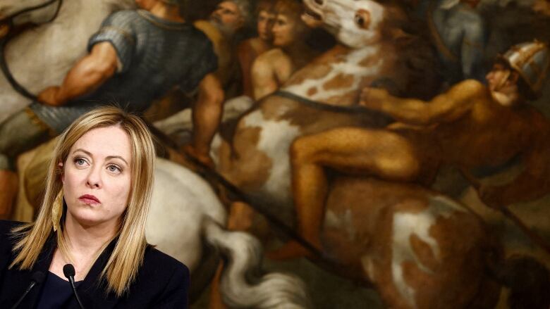 Woman with long blonde hair and serious expression stands in front of a microphone in front of a large mural painting of warriors on horses.