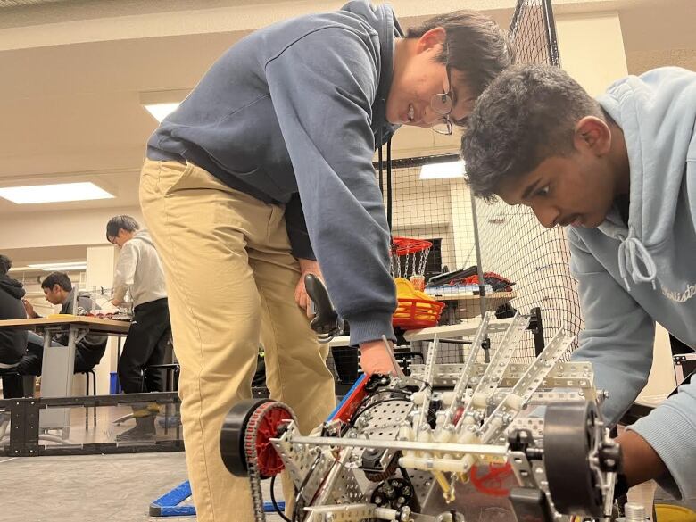 two boys wearing sweaters fix up a robot. the robot has wheels and one of the boys is smiling 