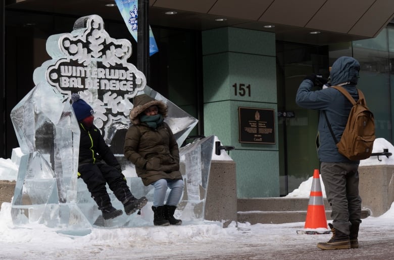 Two people in winter jackets sit on a bench carved out of ice while another person takes their photo.