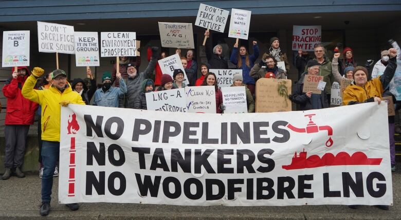 A group of citizens hold banners and signs in front of a public building, protesting Woodfibre LNG.