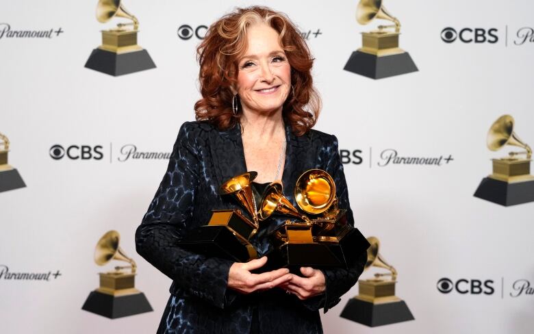 A woman with long, wavy red hair smiled at the camera while holding three brass-coloured awards