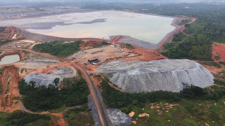 An aerial photo shows a large pond with a road and parking lot leading toward it, surrounded by reddish soil and deep green forests.