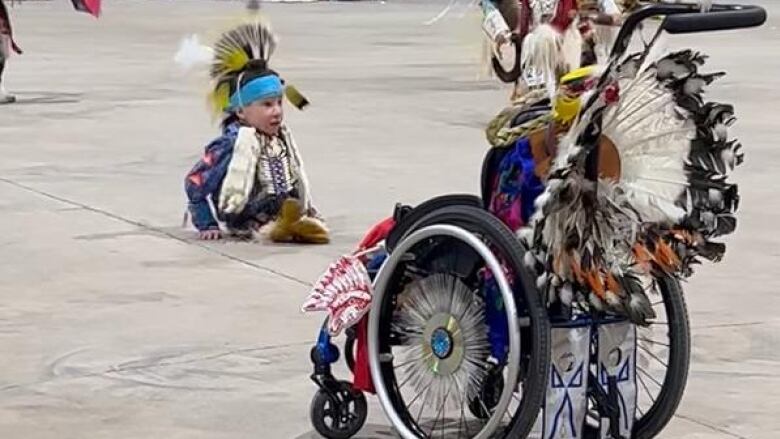 A boy in powwow regalia sitting on the floor with his wheelchair beside him.