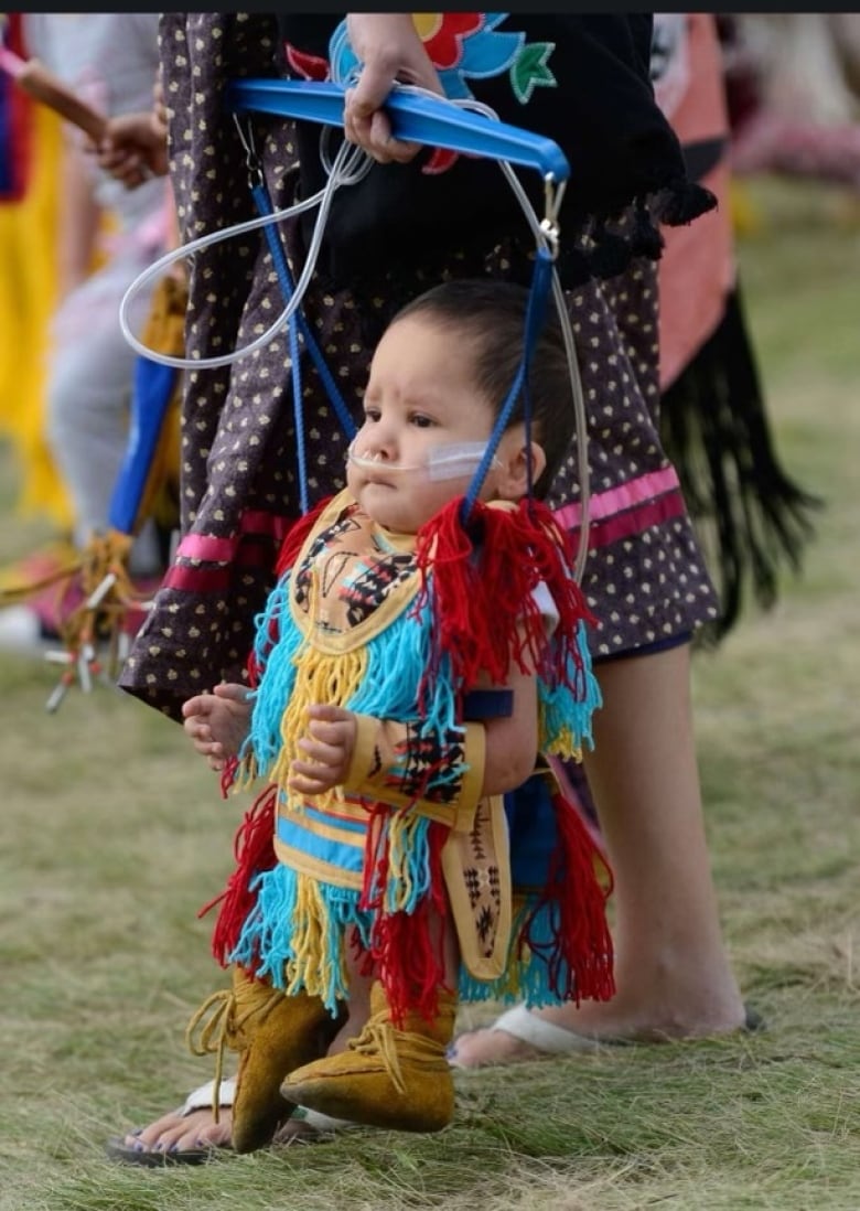 A baby dressed in powwow regalia in a jolly jumper held by his mom.