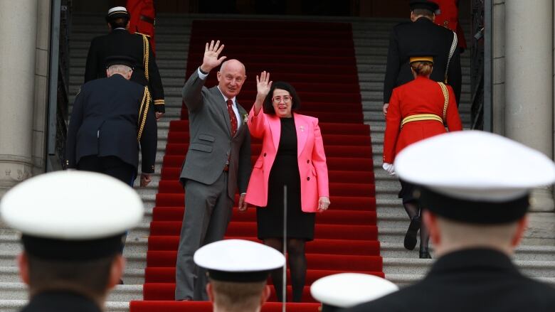 A smiling bald man in a grey suit stands beside a smiling woman with a dark bob hair style and a pink blazer over a black, knee-length dress. They are both waving to the camera on the steps on the B.C. legislature.