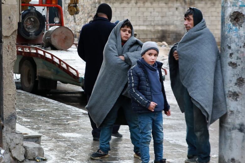 A man and two boys stand on a sidewalk, dusted with snow. The man, on the right, and the boy, on the left are wrapped in thick, grey blankets. A younger boy stands in between, wearing a thick jacket, a hoodie and a hat on his head.