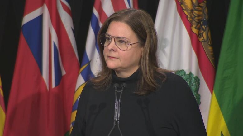 Manitoba's premier, dressed in black and wearing aviator-style glasses, standing at a microphine. A row of provincial flags is behind her.