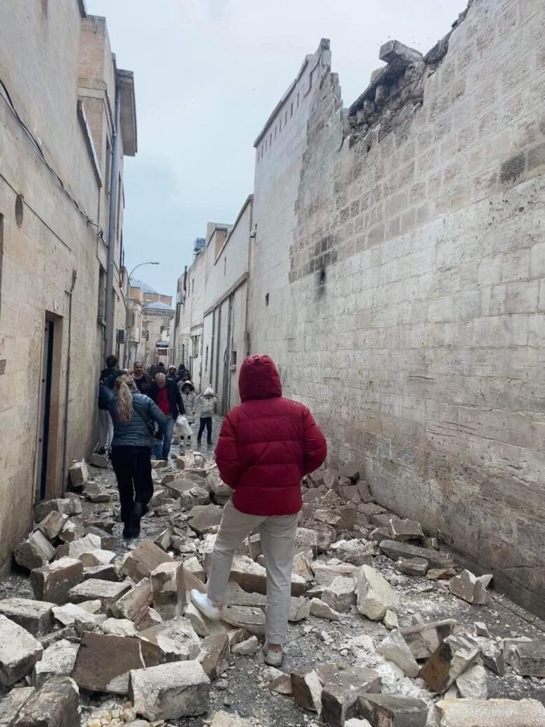 A man in a red jacket walks over fallen stones in a city street. 