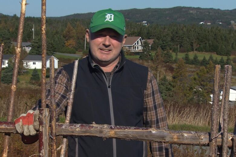 A man wearing a green ball cap and a blue fleece vest rests a gloved hand on a traditional wooden stick fence. 