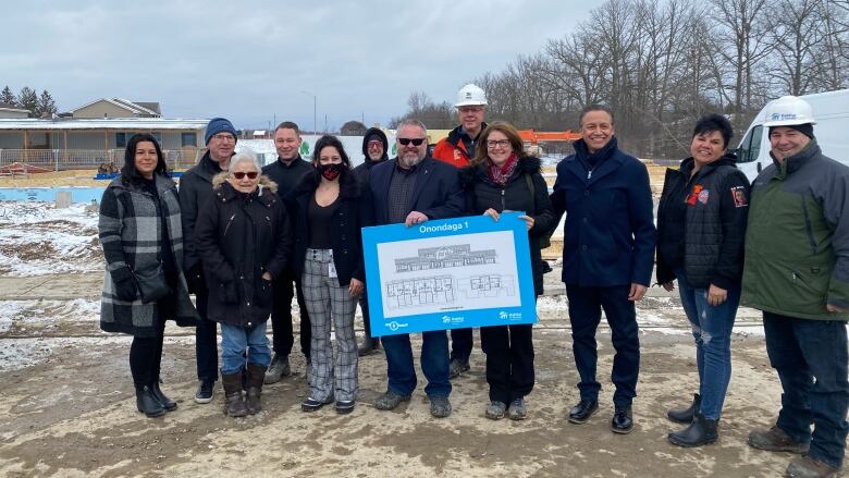 Several people line up outside with a sign, with a construction site and a bit of snow behind them. 