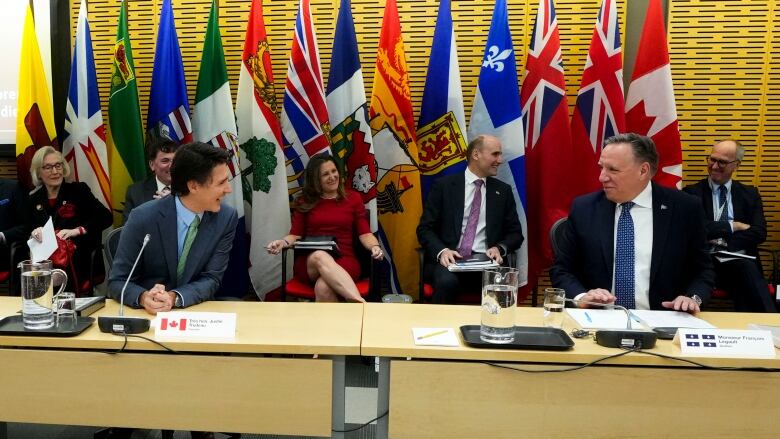 Two smiling men wearing suits sit at a long table with microphones, in front of a row of seated people under a wall with large Canada and provincial flags.