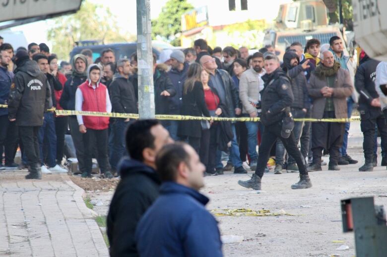 A large group of people gather behind yellow police tape, as a police officer in uniform walks by. To the right of the frame, part of a construction vehicle is shown.