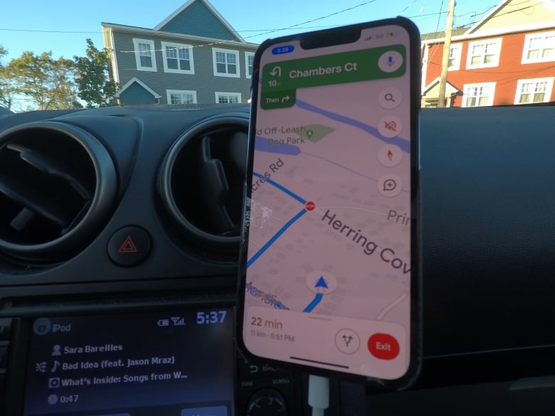 A cell phone is mounted on the dashboard of a car, showing driving navigation directions. Large older residential buildings are seen through the windshield.