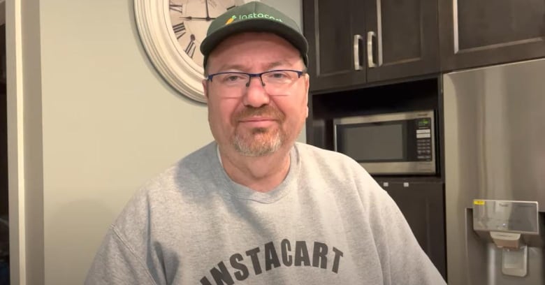 A smiling white man with a short-cropped goatee and glasses is wearing a light grey Instacart sweatshirt and green Instacart-branded baseball hat. He is standing in a kitchen with a wall clock, cupboards, and a microwave oven in the background.