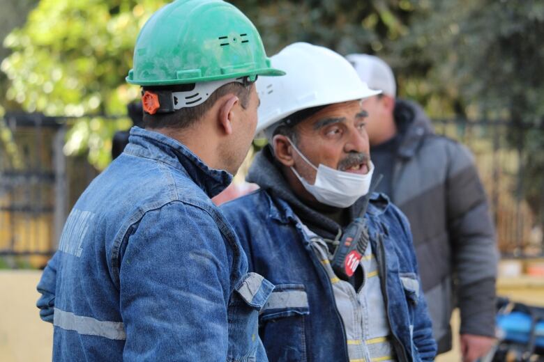 Two men wearing blue jumpsuits and hard hats talk to each other. One of them is wearing a face mask that's pulled just below his mouth.