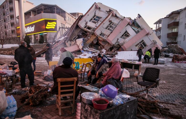 A group of people sit on chairs on the street. A pink building has collapsed behind them.