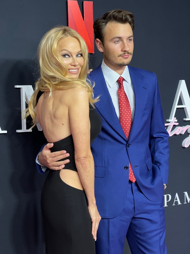 A woman in a black dress smiles and stands next to a man wearing a blue suit and a red tie.