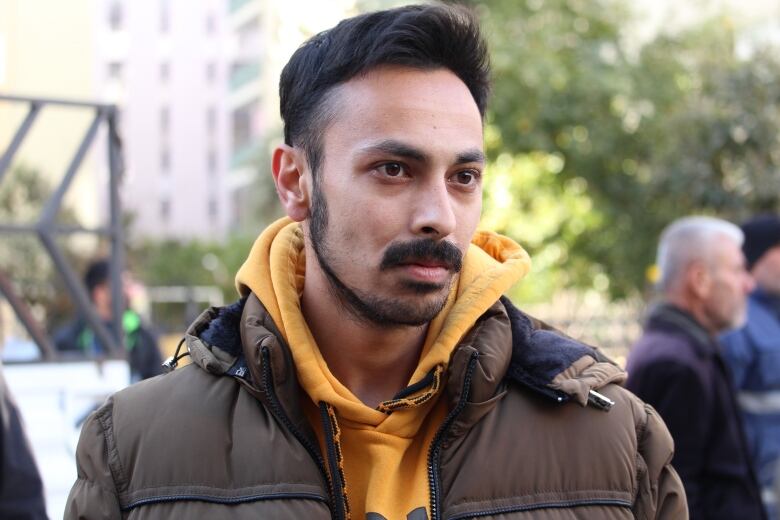 A man with a mustache and beard, wearing a yellow hoodie with a brown jacket over top, is shown outside. Buildings, trees and a couple of other people are blurred in the background behind him.