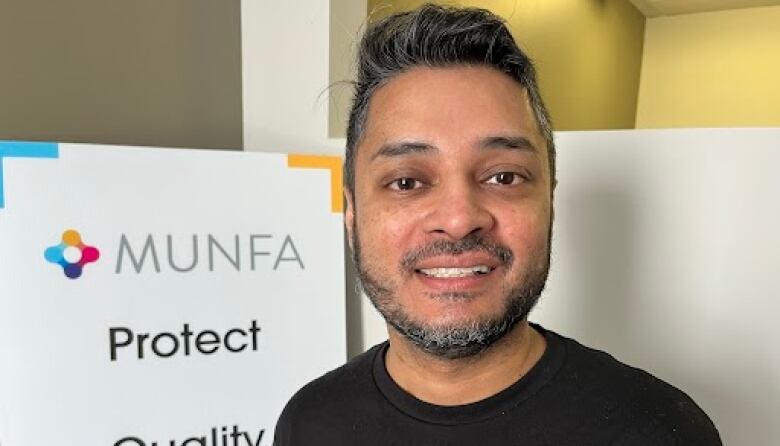 A man with a greying beard smiles at the camera, with a sign behind him that says MUNFA.