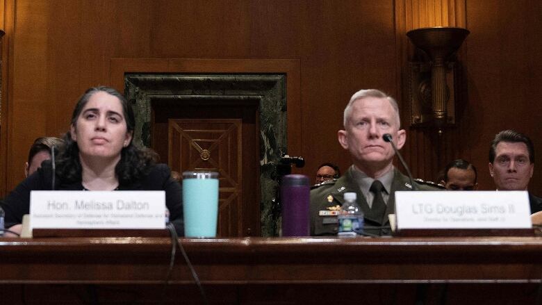 A woman and man are shown behind microphones at a long table in a wood-paneled room.