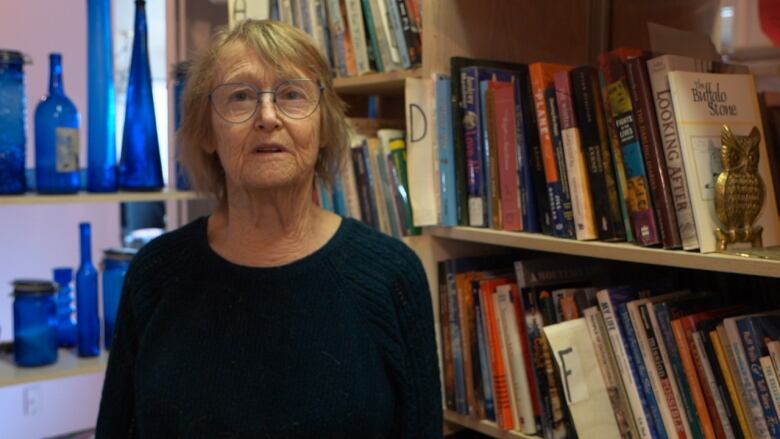 A woman wearing a black sweater stands in front of book shelves and a shelf of blue glass bottles and vases.