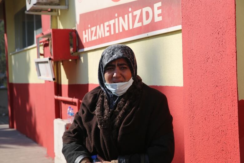 A woman wearing a grey headscarf with a light swirl pattern and a brown jacket stands outside a building with yellow and red plastered walls. She's also wearing a face mask pulled down just below her mouth.