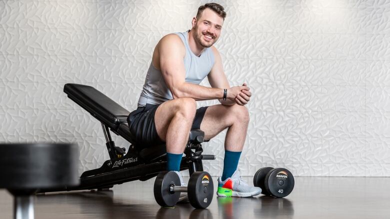 A middle-aged man sits on a workout bench wearing black shorts and a tank top. He smiles at the camera for this professional photo.