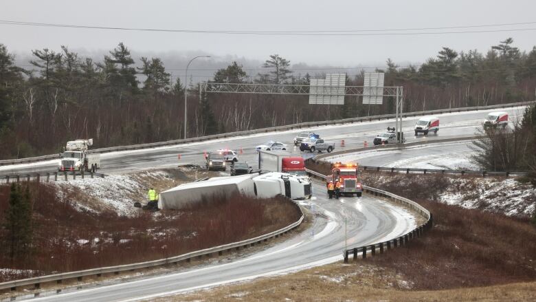 A transport truck lies in a grassy area alongside an exit ramp from a highway as emergency vehicles sit nearby with their lights flashing.