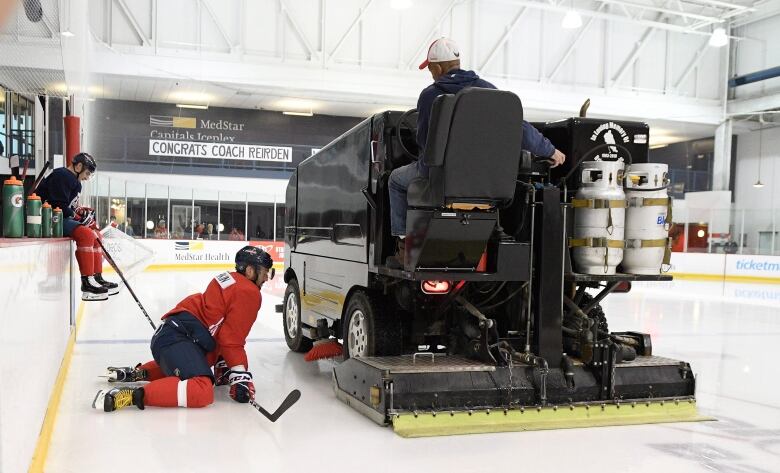 An ice resurfacer with two gas tanks passes beside a hockey player kneeling on the ice.