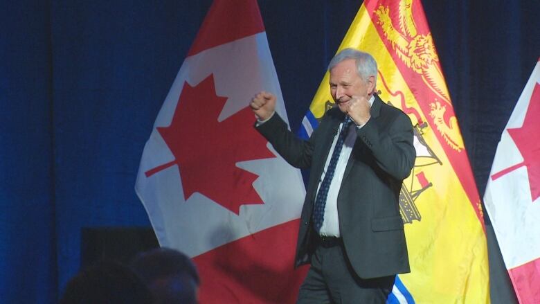 A man with white hair in a dark suit raises his hands while he dances on stage.