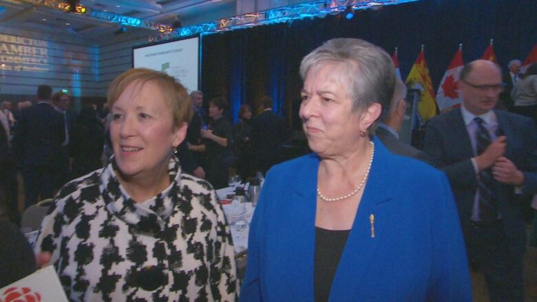 Two woman, one in black and white checks, one in blue, stand in a banquet room.