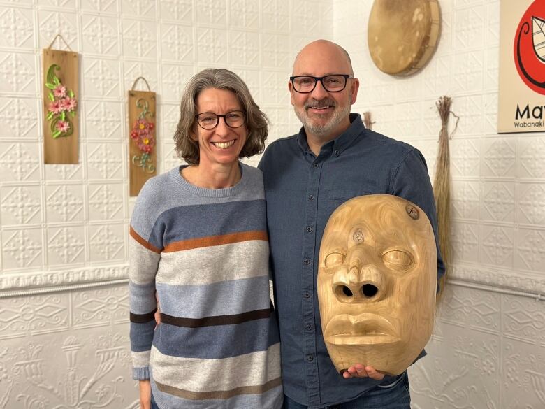 A woman and man stand with their arms around each other. The man holds a large, wooden mask.