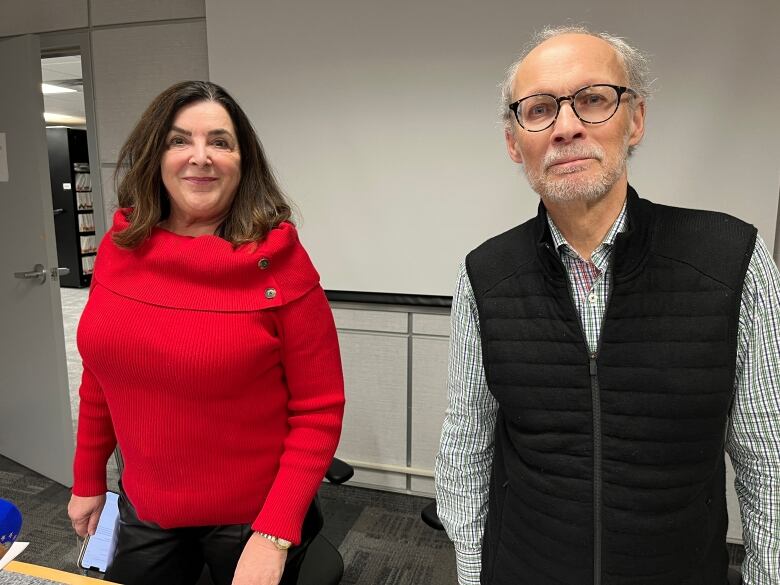 A woman wearing red and a man wearing a white shirt with a black vest stand inside a board room. Behind them is a blank projector screen. 