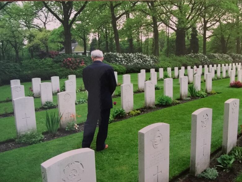 An elderly man, back turned to the camera, surveys a group of graves in a military cemetery.