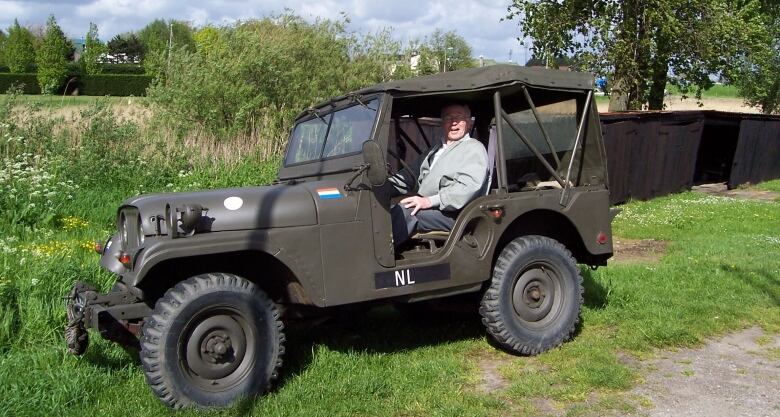 An elderly man sits in a military jeep, smiling at the camera.