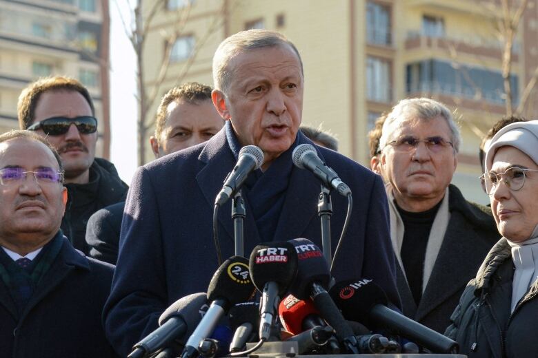 A man stands at a clusters of microphones, with eight people standing around him. Two multi-storey buildings are visible in the background.