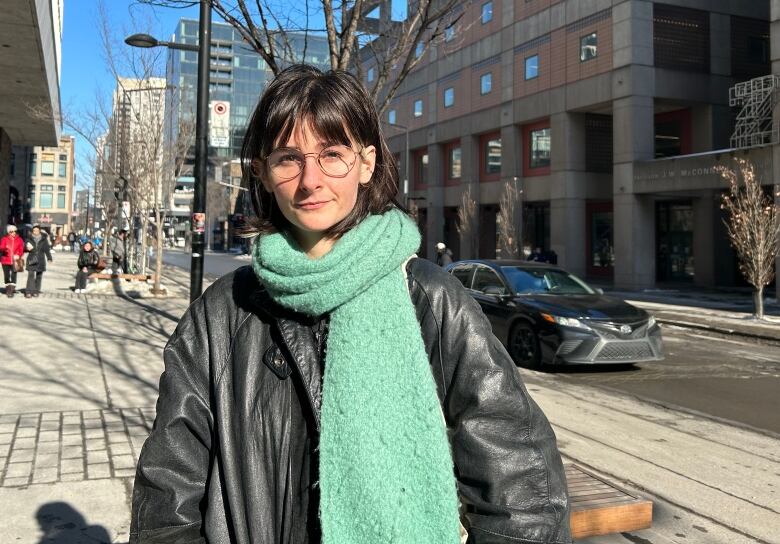 A brunette wearing a leather jacket, green scarf and round glasses looking at the camera in front of Concordia University.