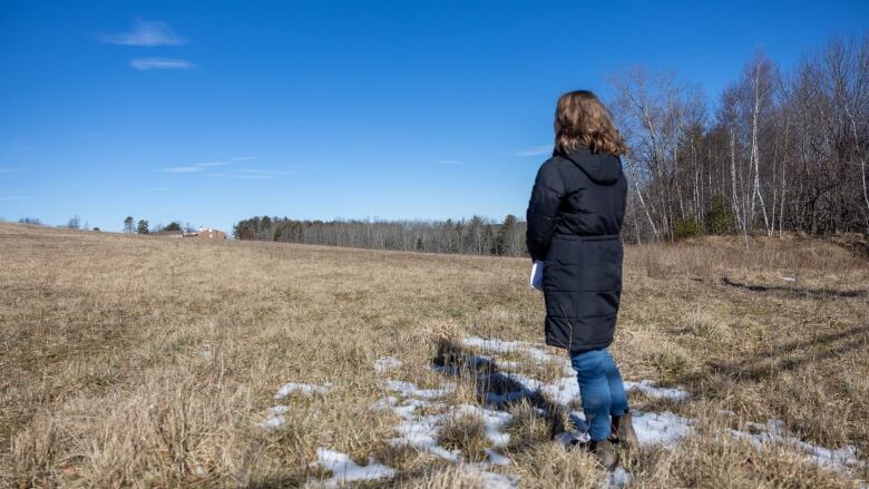 A woman with brown hair in a black coat stands facing away from the camera toward a line of trees, with clear blue sky above.