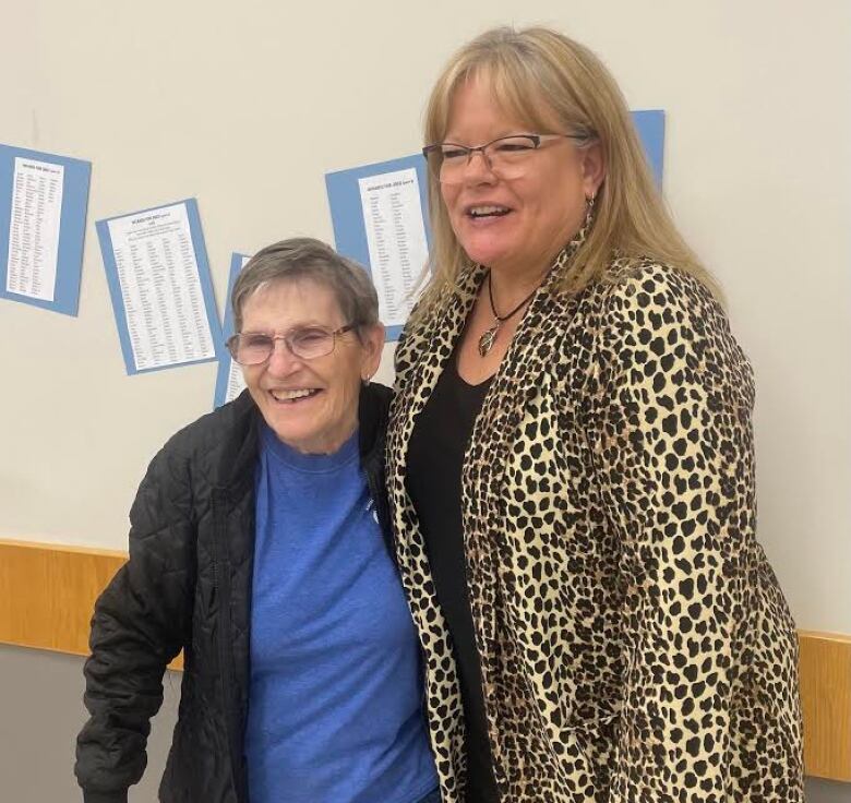 Two women stand with their arms over each other's shoulders and smile.