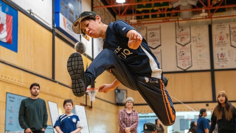 A boy leaps into the air, one foot back, the other forward in an effort to hit a ball hanging from a rope with his foot.