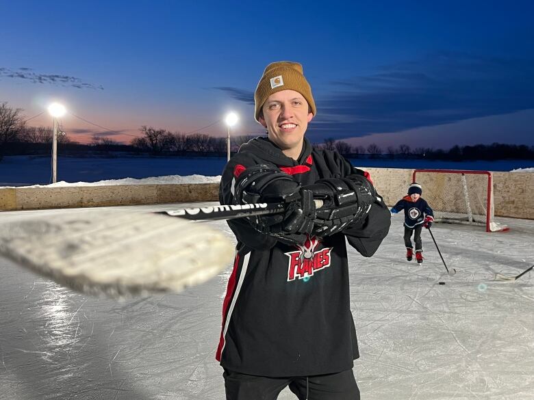 A man stands holding a hockey stick at an outdoor rink.