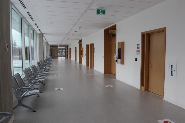 A long hallway with a row of chairs next to a window on one side and wooden doors on the other.