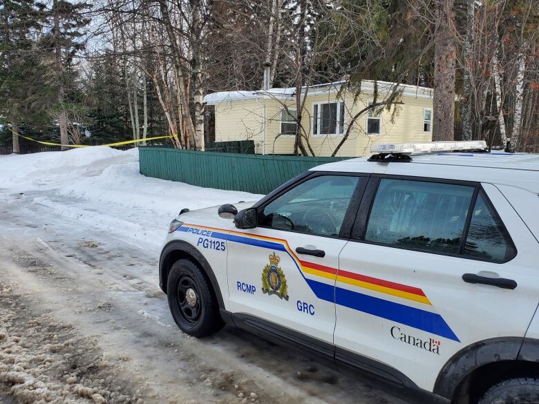 An RCMP cruiser sits on a snowy road leading to a yellow house surrounded by trees.