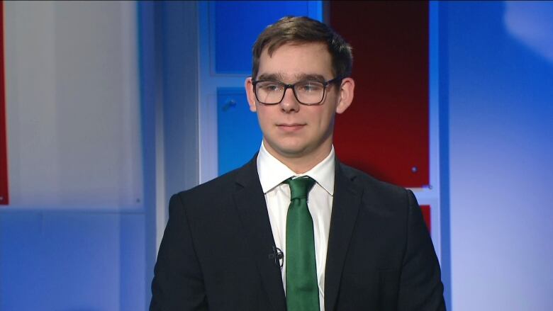 A man in a blazer and green necktie sits in a TV studio.