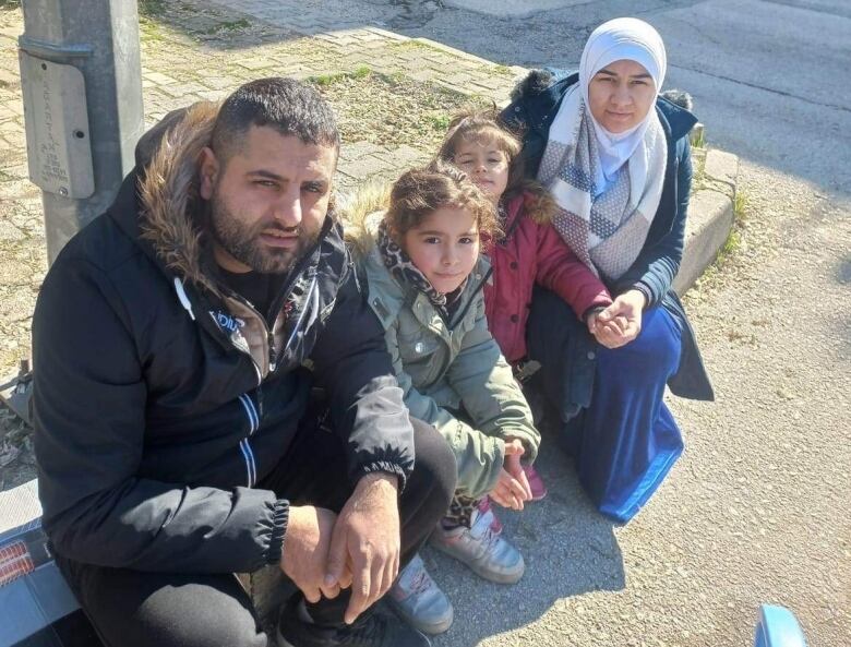 A couple sits outside on on a curb with their two young daughters. 