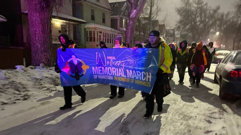 People dressed in winter clothing walk on a snow-covered residential street in the dark, carrying a blue banner that says Annual Women's Memorial March of Manitoba in honour of MMIWG2S. 