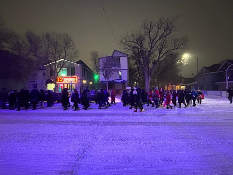 A wide shot of several people marching on a snow-covered street with buildings lit up in the background.