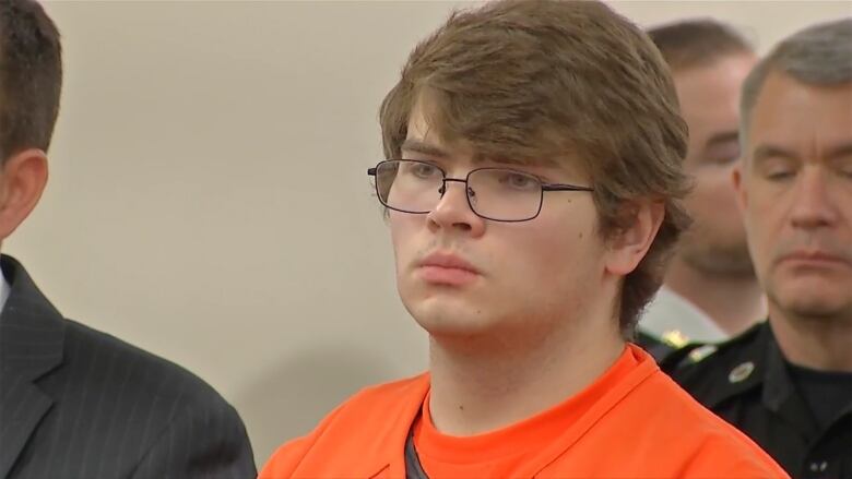 A young man in glasses in prison garb is shown standing.