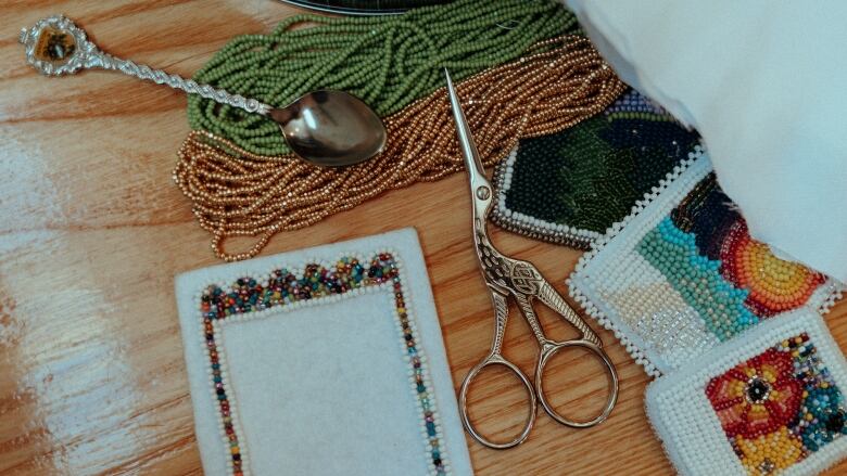 An unfinished square of beadwork lies on a table near some finished pieces, beads and other equipment.