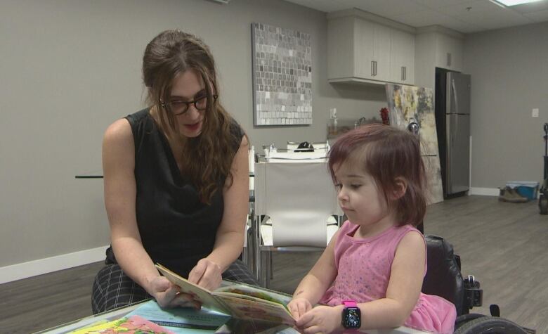 A woman in a black top and plaid trousers reads a book to a little girl dress in a pink dress sitting on a wheelchair next to her.